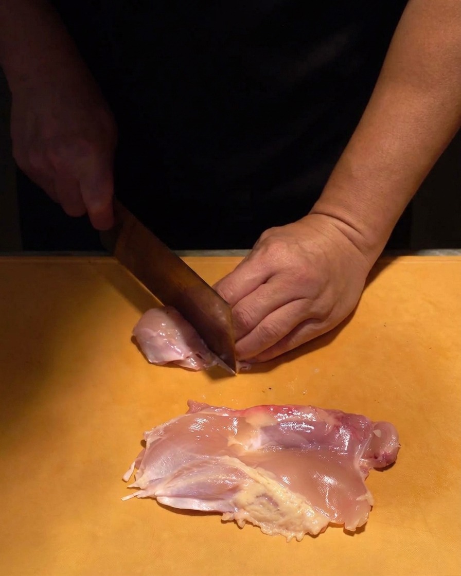 オレンジ色のまな板の上で、生の鶏もも肉を一口大に切っている様子。
