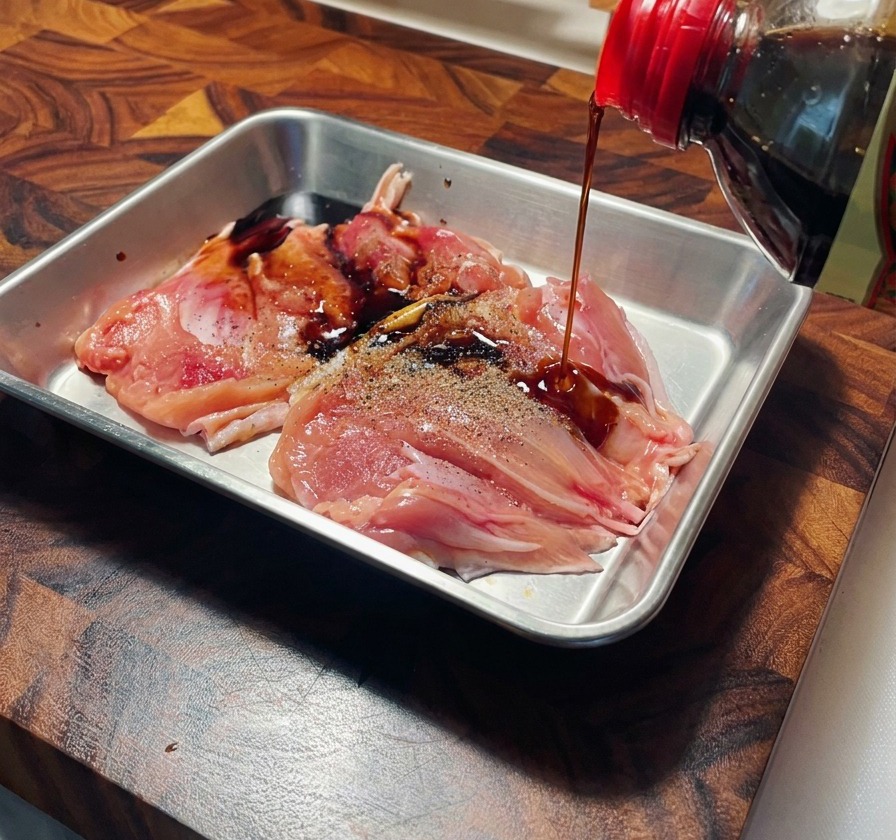 ステンレスバットの上で、胡椒を振った鶏もも肉に醤油をかけている様子。