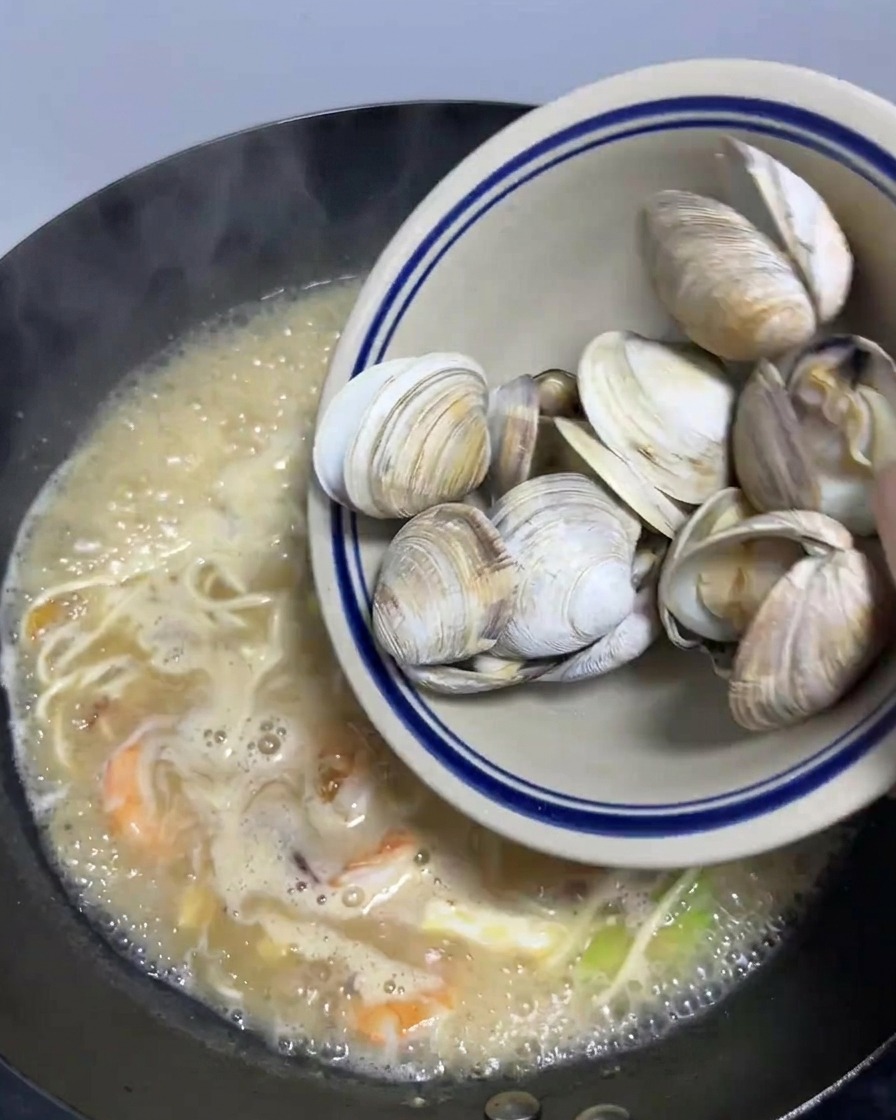 煮え立った海鮮ラーメンの鍋に、下ゆで済みのあさりを投入している様子。