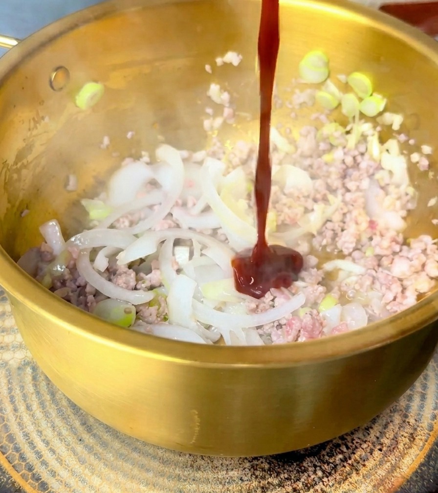 炒めた豚肉と玉ねぎの上に、濃い赤色のビビンバソースを注いでいる様子。