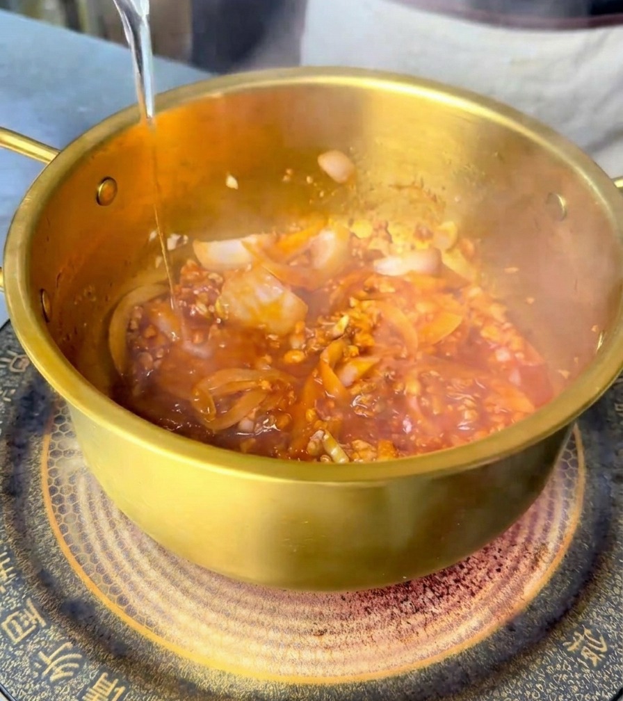 赤いスパイスベースが入った鍋に水を注ぎ、混ぜ合わせている様子。