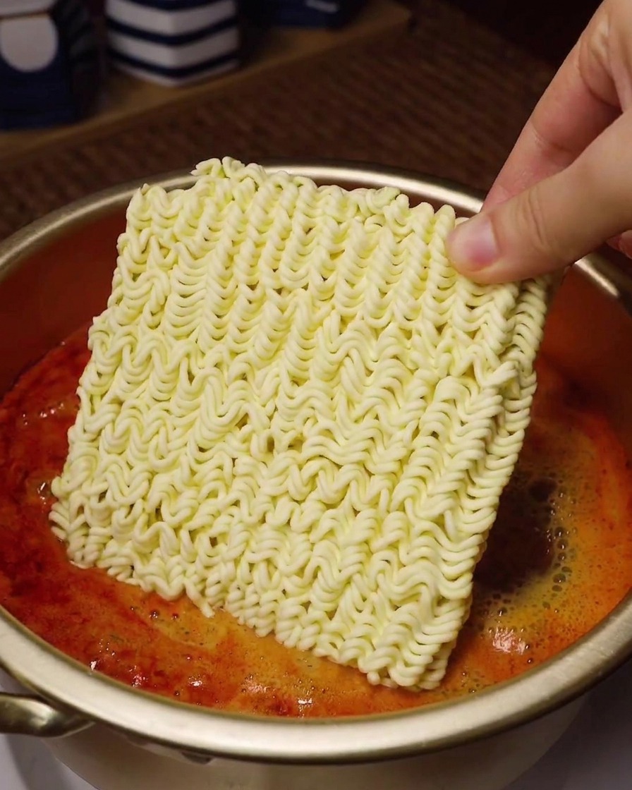 ぐつぐつと煮立つ真っ赤なスープが入った金色の鍋の上で、四角いインスタントラーメンの乾麺を手に持っている様子。