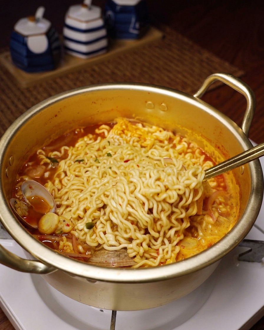 ぐつぐつと煮える赤い海鮮スープから、ちぢれ麺を金属製のスプーンですくい上げている様子。