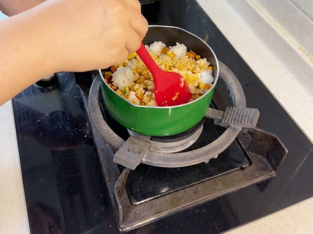 コンロの上の緑色のフライパンで、赤いヘラを使って白いご飯と炒めた鮭や野菜を混ぜ合わせている様子
