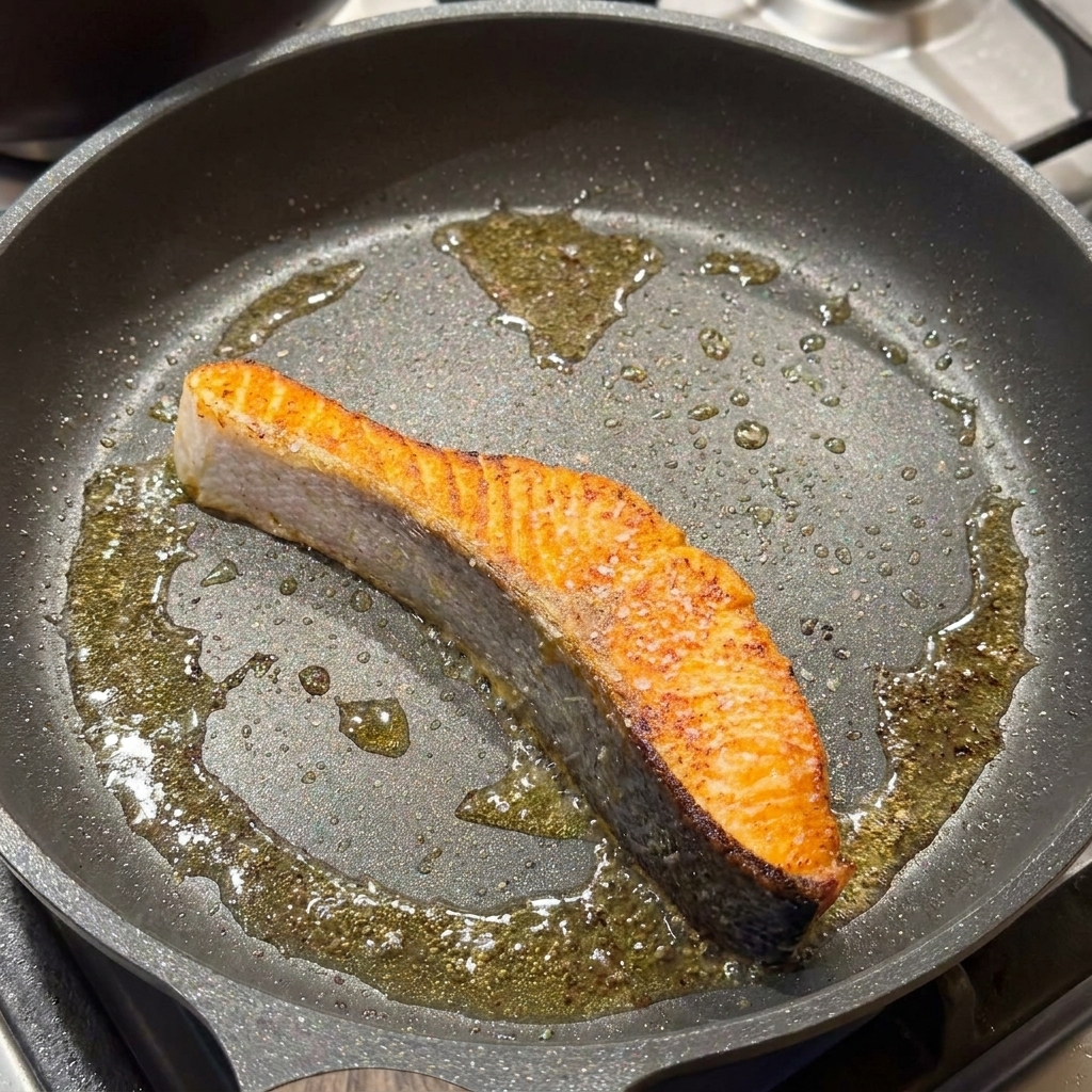フライパンで焼いている鮭に塩と黒こしょうを振っている様子。