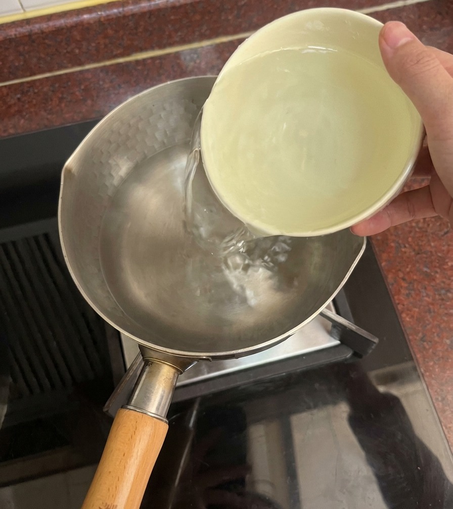コンロに置かれた鍋に、陶器のボウルから透き通った水を注いでいる様子。