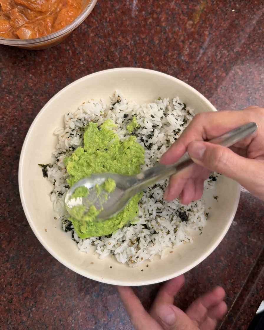 白いボウルに入った温かいご飯と刻みのりの上に、鮮やかな緑色のアボカドをスプーンで広げている様子。