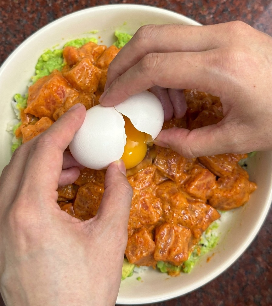 サーモンとアボカドが盛られた丼の中央で、白い卵の殻を割っている様子。