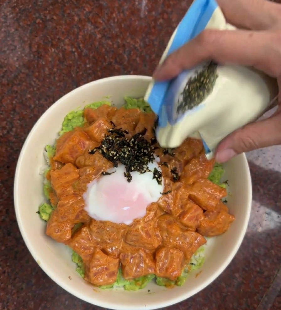 青と白の小袋から、刻みのりとごまのミックスをサーモン丼に振りかけている様子。