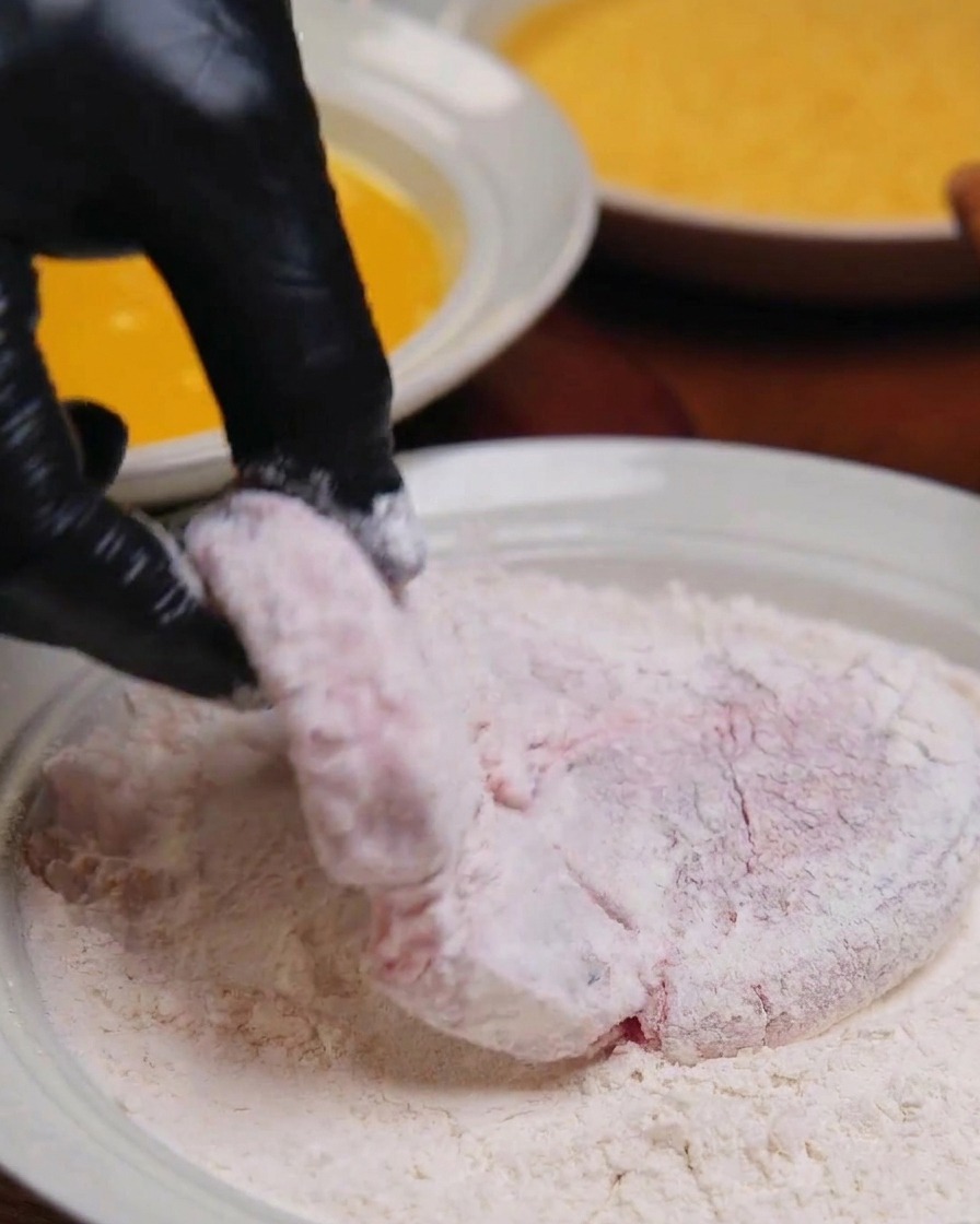 手袋をした手で、小麦粉を全体にまぶした厚切りの豚肉を持っている様子。