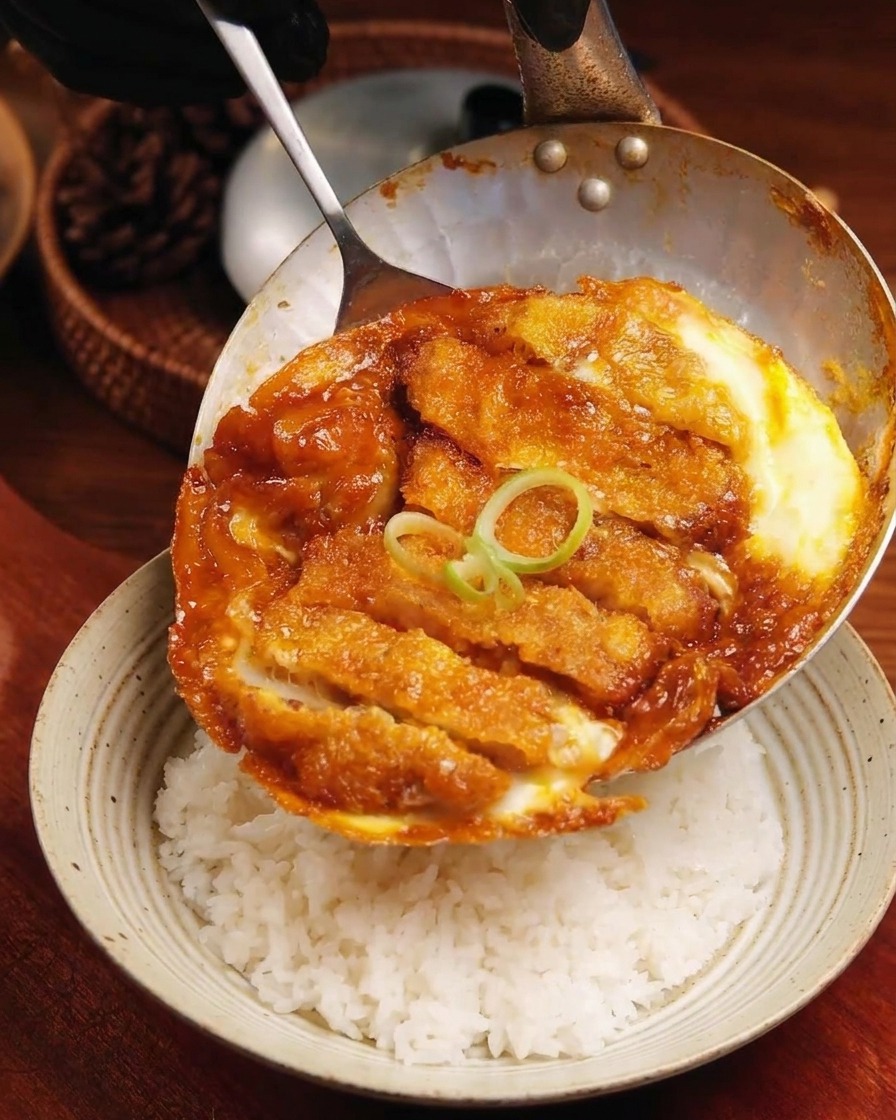 フライパンからカツと卵の混合物を、丼のご飯の上に滑らせるようにのせている様子。
