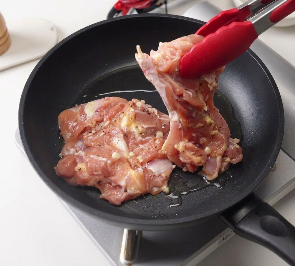 熱した黒いフライパンに、トングで下味をつけた鶏肉を皮目を下にして入れている様子。
