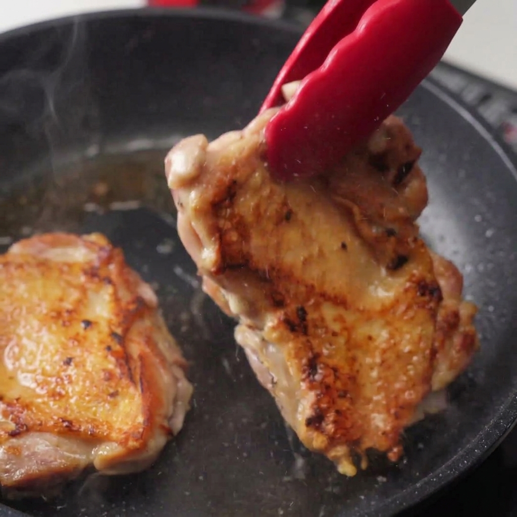 フライパンの中の鶏肉をトングで裏返し、こんがりと焼けた皮目が見えている状態。湯気が立ち上がっている。