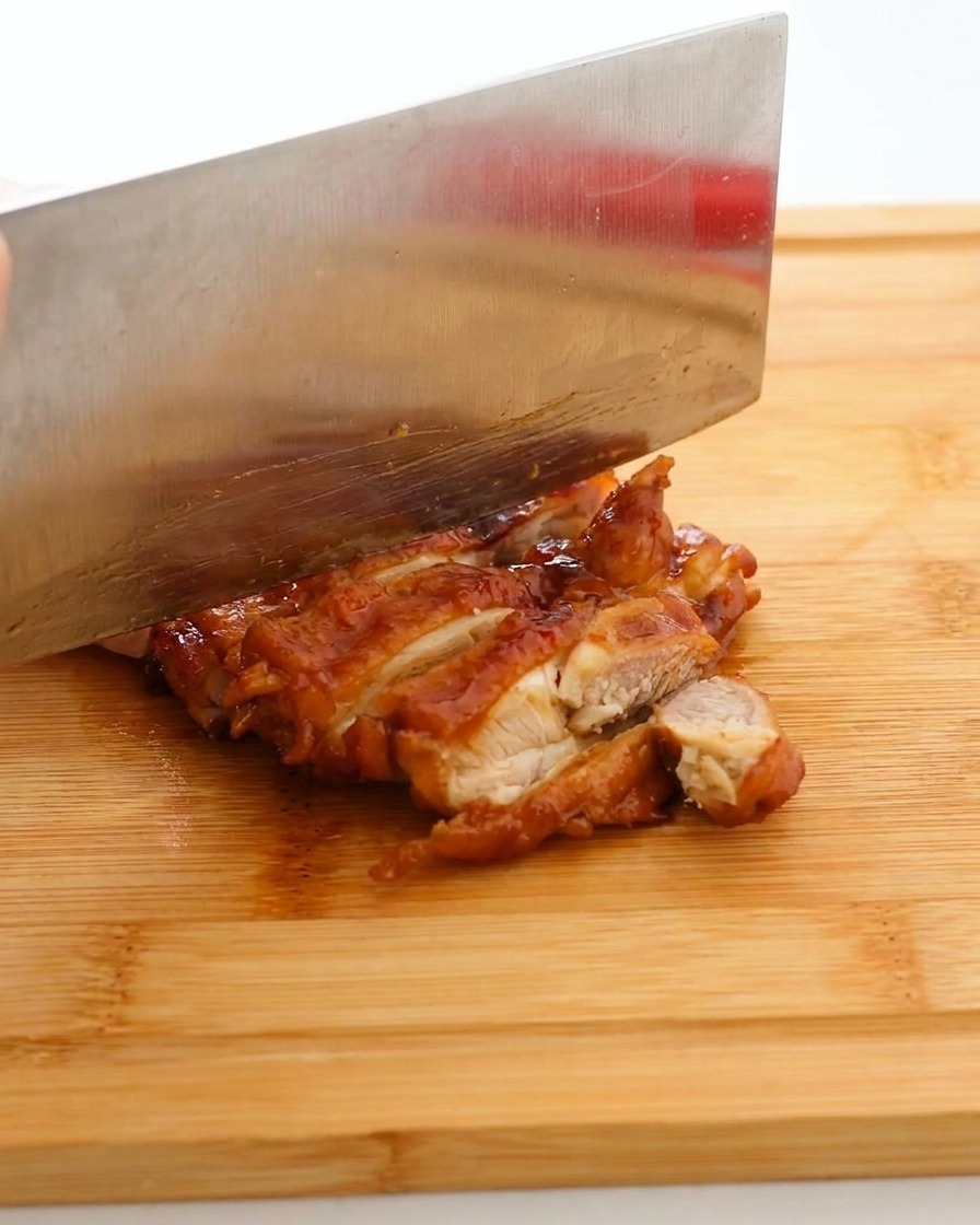 照りよく焼けた鶏肉を、まな板の上で包丁を使って食べやすい大きさに切っている様子。