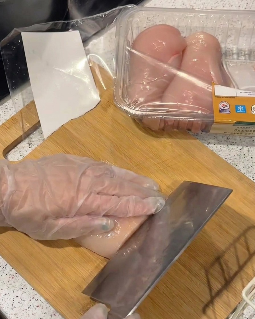 竹のまな板の上で、手袋をした手で生の鶏むね肉を押さえながら、包丁で薄くスライスしている様子