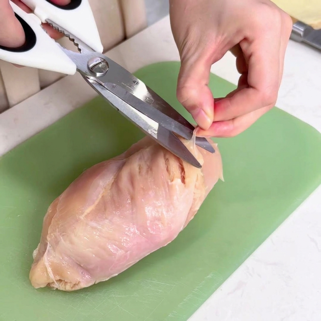 グリーンのまな板の上で、キッチンバサミを使って鶏肉の表面にある白い薄膜を切り取っている様子。