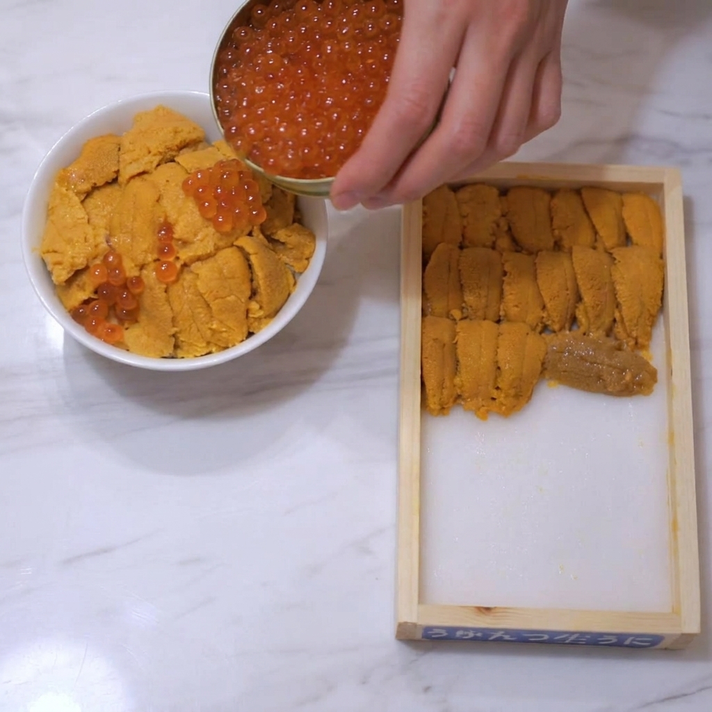 うにが乗った丼の上に、小さな金色の缶から輝くオレンジレッドのいくらを注ぎ入れる様子。