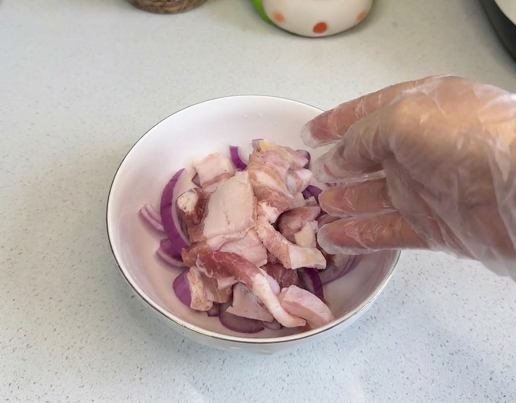 透明なプラスチック手袋をはめた手が、赤玉ねぎの輪切りが入った白いボウルに生の豚バラ肉のスライスをのせている様子。