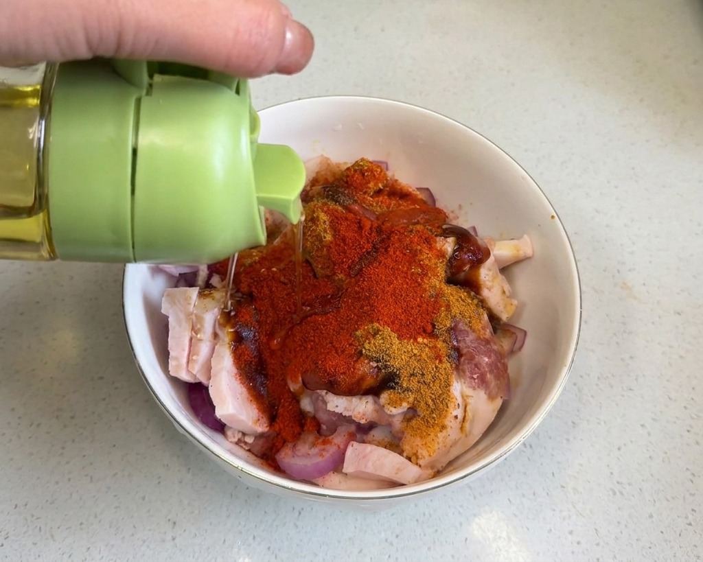 赤いスパイスで覆われた豚バラ肉に、緑色のキャップの容器から透明な調理油が注がれている様子。