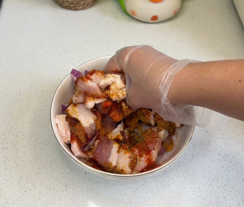 手袋をはめた手が、白いボウルの中で生の豚バラ肉、玉ねぎ、調味料を徹底的に混ぜ合わせている様子。