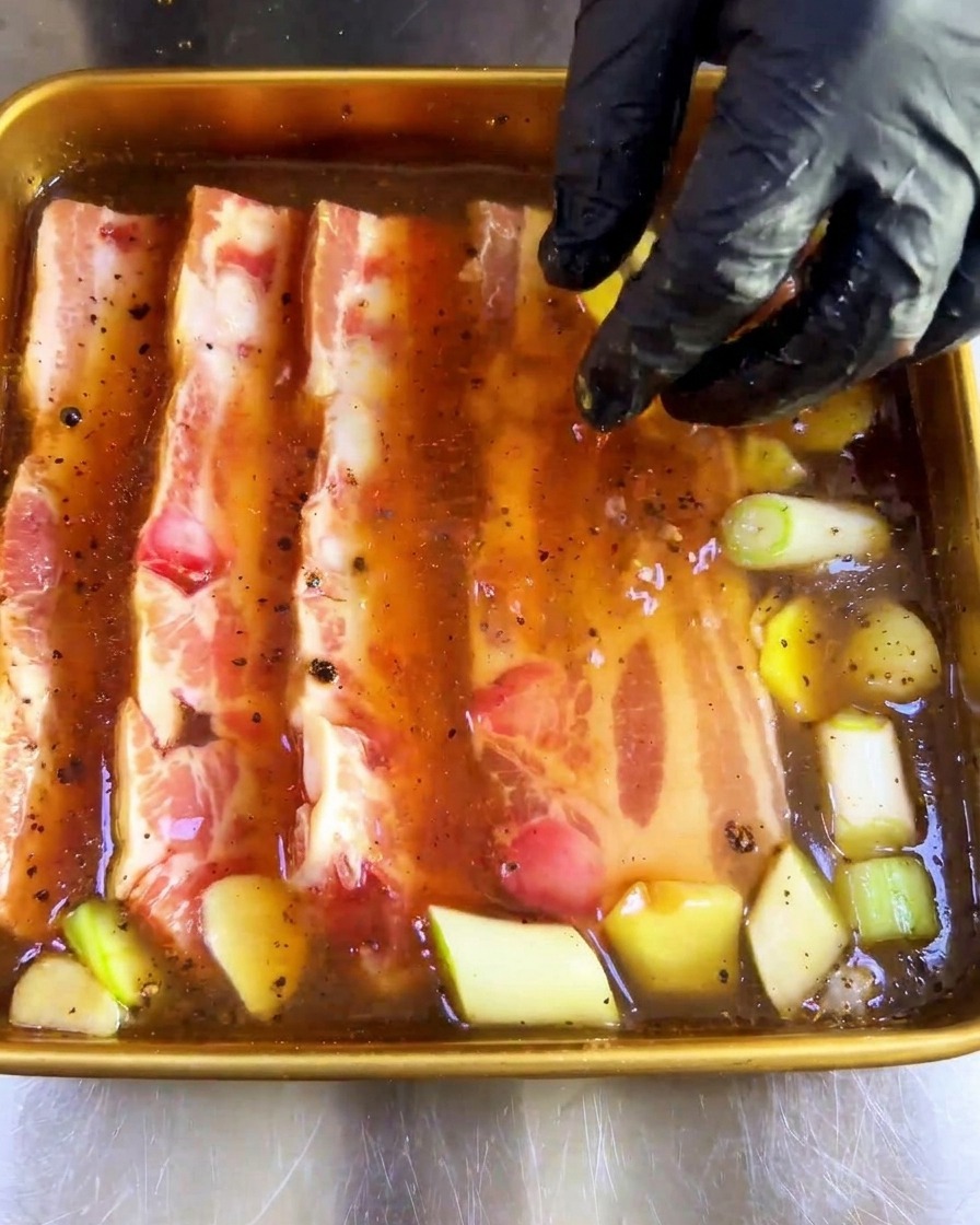 手袋をした手で、厚切りの豚バラ肉をタレの中に沈めている様子。