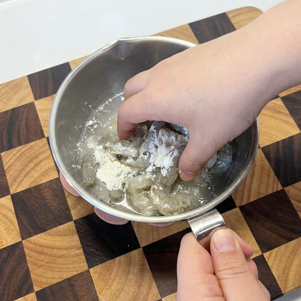 金属製のボウルで、生の海老と白い片栗粉を手で混ぜ合わせている様子