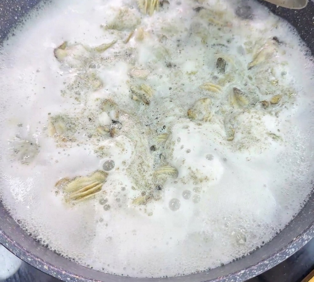 激しく沸騰し白い泡が立っている鍋から、網杓子で茹でた牡蠣をすくい上げている様子。