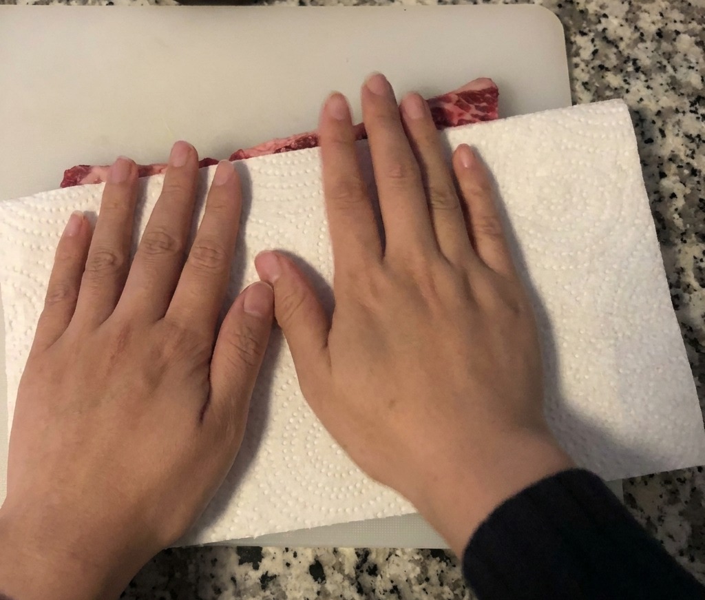 白いまな板の上に置かれた生の骨付き牛カルビ肉を、白いキッチンペーパーで押さえて水気を拭き取っている様子。