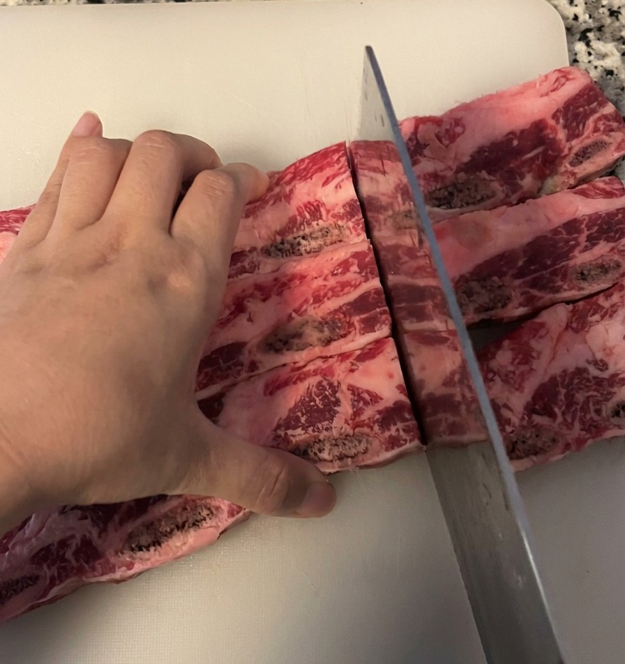 白いまな板の上で、大きな包丁を使って生の骨付き牛カルビ肉を骨の間で切り分けている様子。