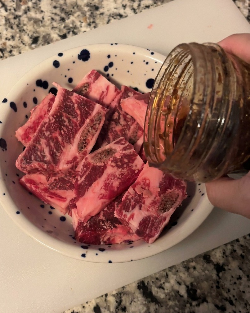 青い模様の入った白いボウルに入った生のカット済み牛カルビ肉に、ガラス瓶から濃い色でツヤのある韓国風カルビソースを注いでいる様子。