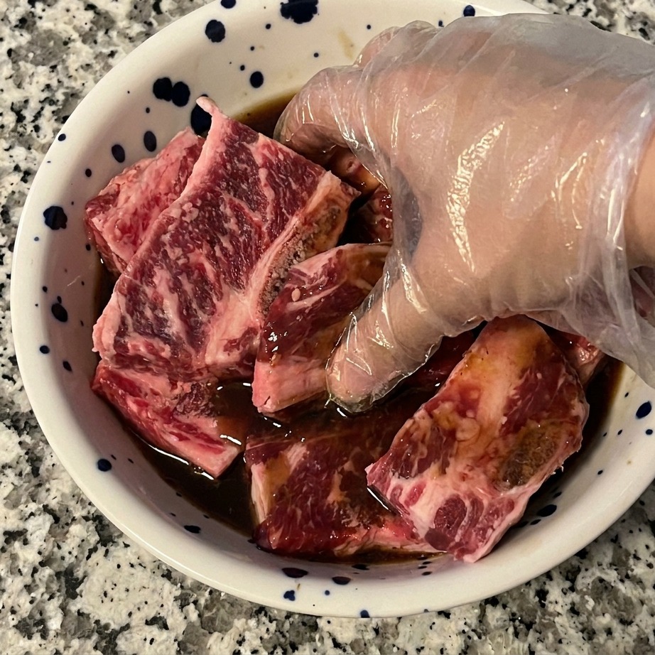 透明なビニール手袋をした手で、生の牛カルビ肉と濃い色のマリネ液をボウルの中で揉み込み、均一に絡ませている様子。