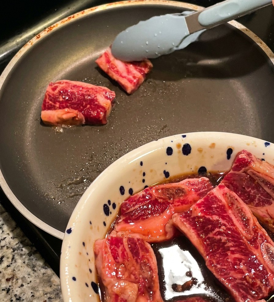 大きなフライパンに薄く油を引き、トングを使って漬け込んだ厚切りの生牛カルビ肉を並べている様子。手前にはお肉の入ったボウルが見える。