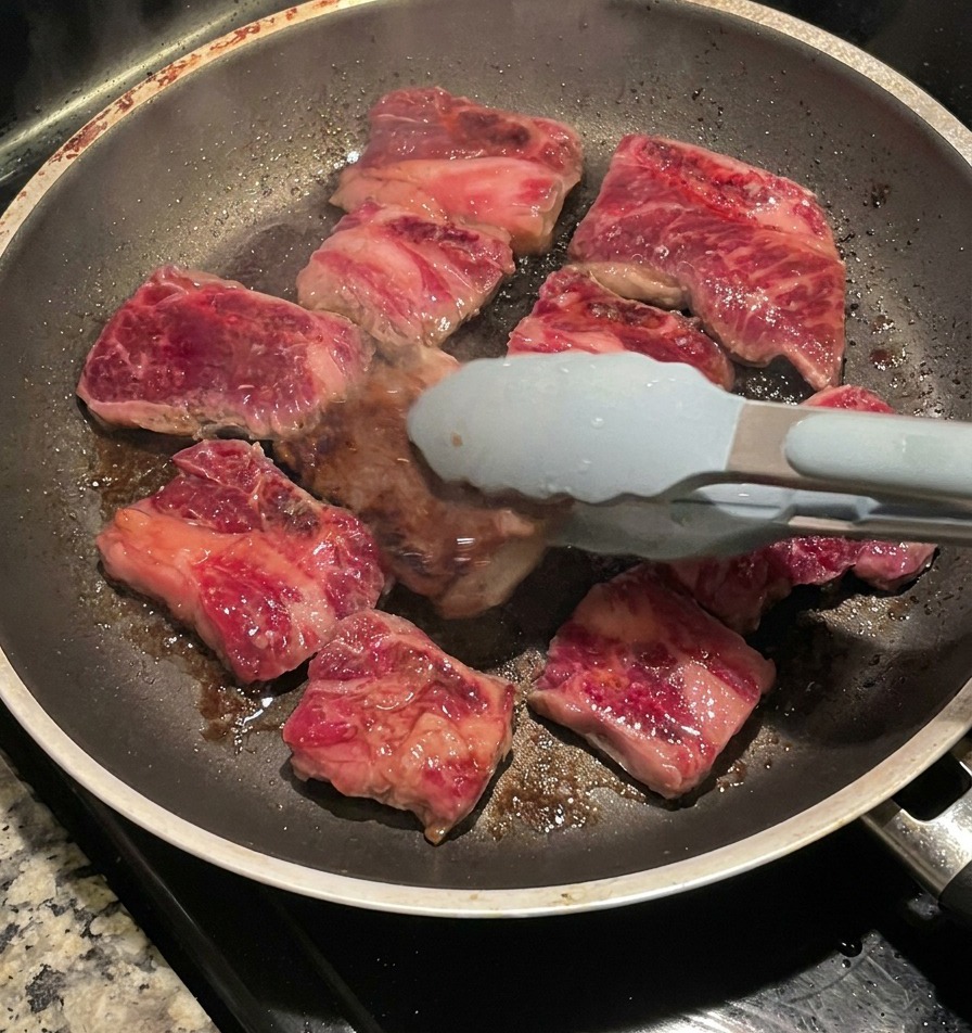青い先端のトングを使って、フライパンで焼いた牛カルビ肉を裏返し、こんがりとキャラメリゼされた焼き色を見せている様子。