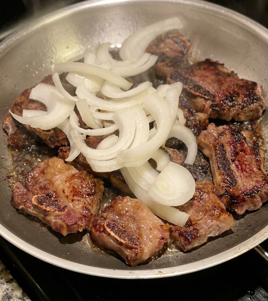 ステンレス製のフライパンの中で、こんがりと焼けた牛カルビ肉の上に、生のスライス玉ねぎがこんもりと乗せられている様子。