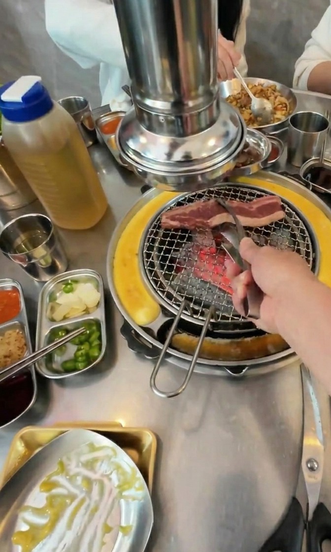 炭火の網の上に、脂ののった長い牛バラ肉を並べている様子。周りには韓国の副菜が並んでいる。