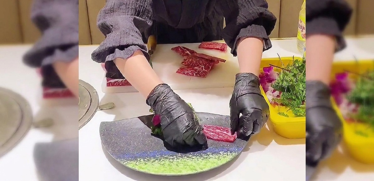 黒い手袋をはめた手が、濃い色の皿の上にスライスした霜降り肉を扇形に重ねながら丁寧に並べている様子。