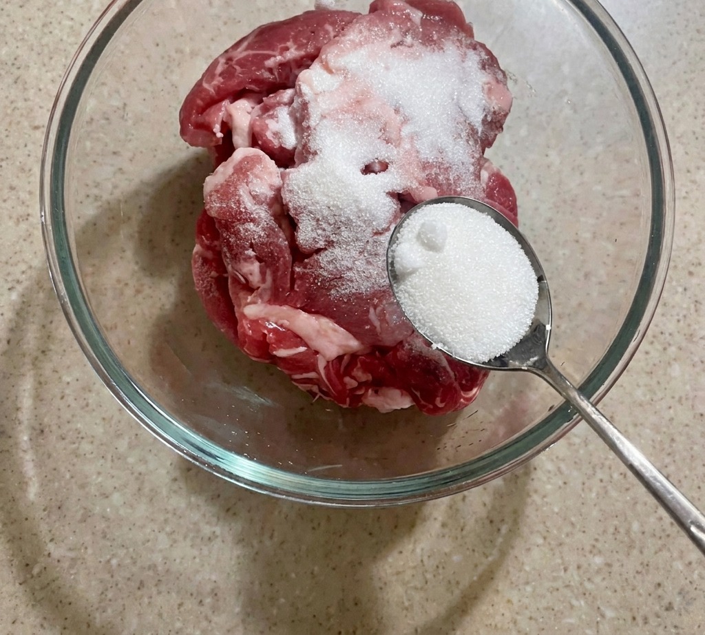 ボウルに入れた薄切りの豚肉に、スプーン一杯の白い砂糖を加えている様子。