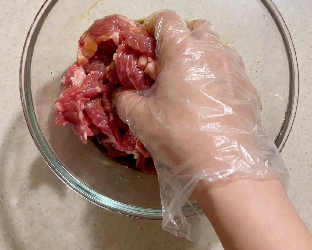 ビニール手袋をはめた手で、ガラスボウルの中の豚肉と調味料をしっかり混ぜ合わせている様子。