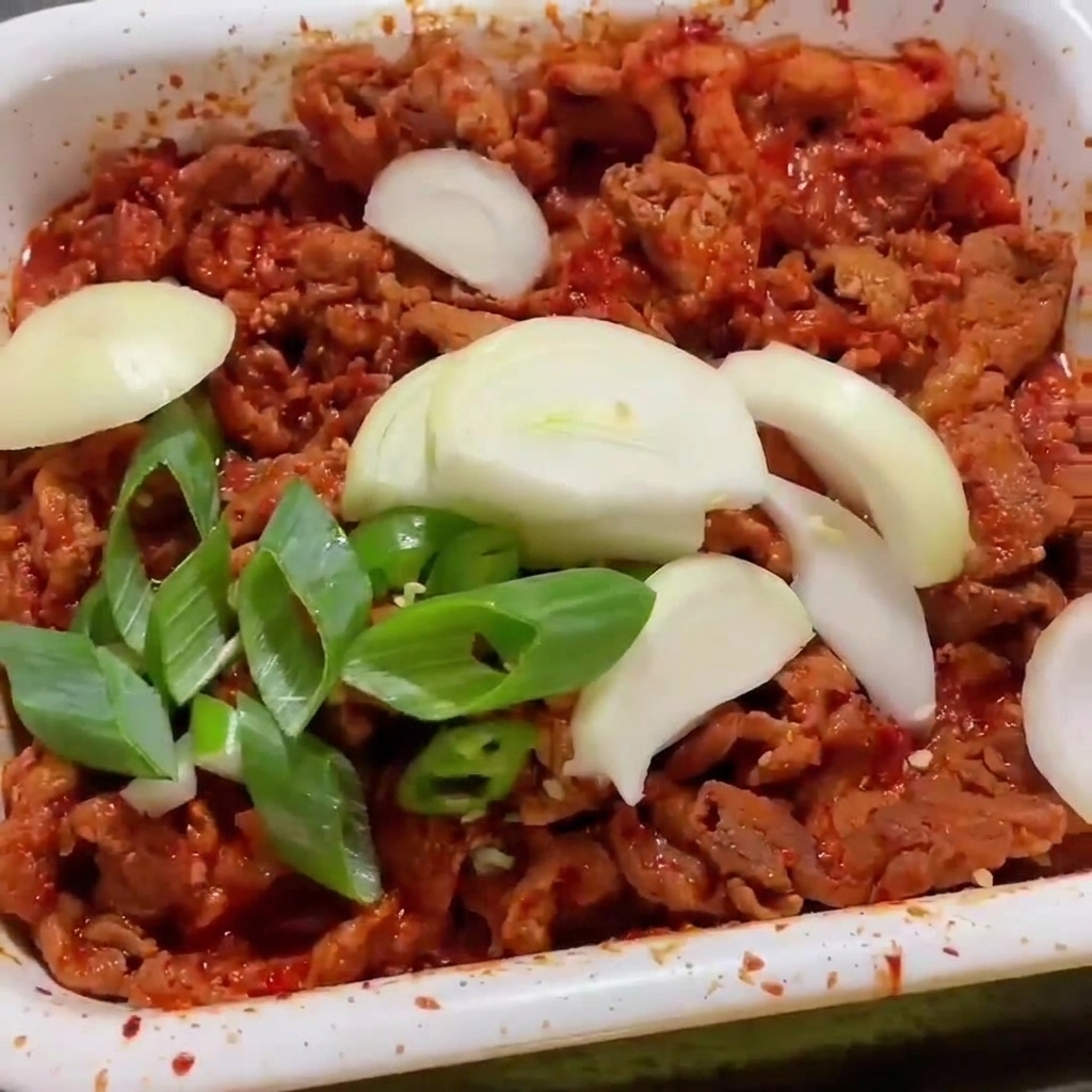 赤いタレが絡んだ豚肉の上に、スライスした玉ねぎと長ねぎをのせて炒めている様子。