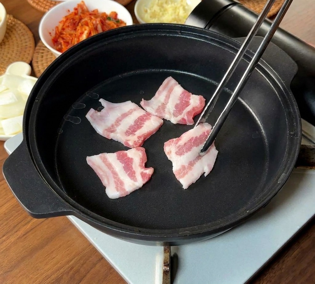 霜降りの豚バラ肉の薄切りを、長い金属製の箸で黒いフライパンに並べている様子。