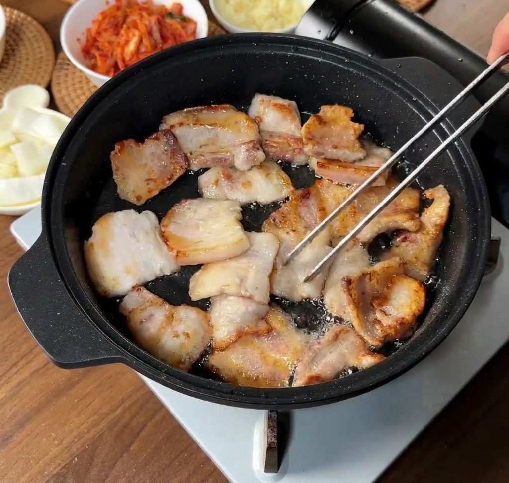 黒いフライパンで豚バラ肉がこんがりと焼け、トングでひっくり返している様子。