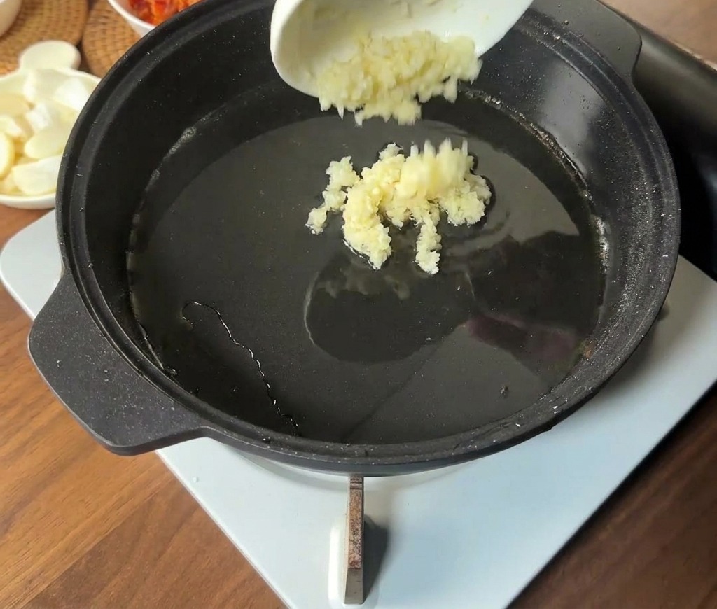 小さな白い器からみじん切りのにんにくを、油の出た熱いフライパンに投入している様子。