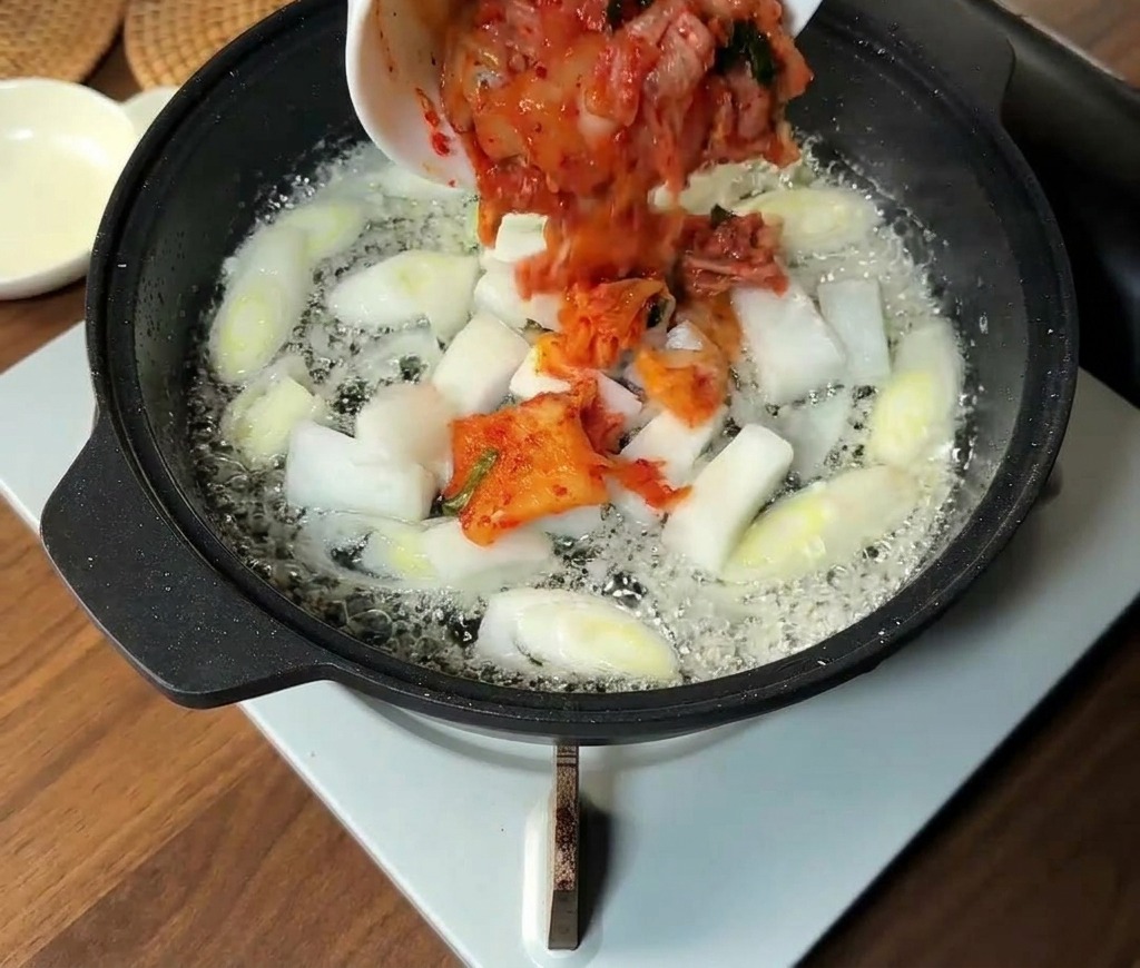 刻んだキムチと玉ねぎのスライスを、野菜を炒めているフライパンに加えている様子。