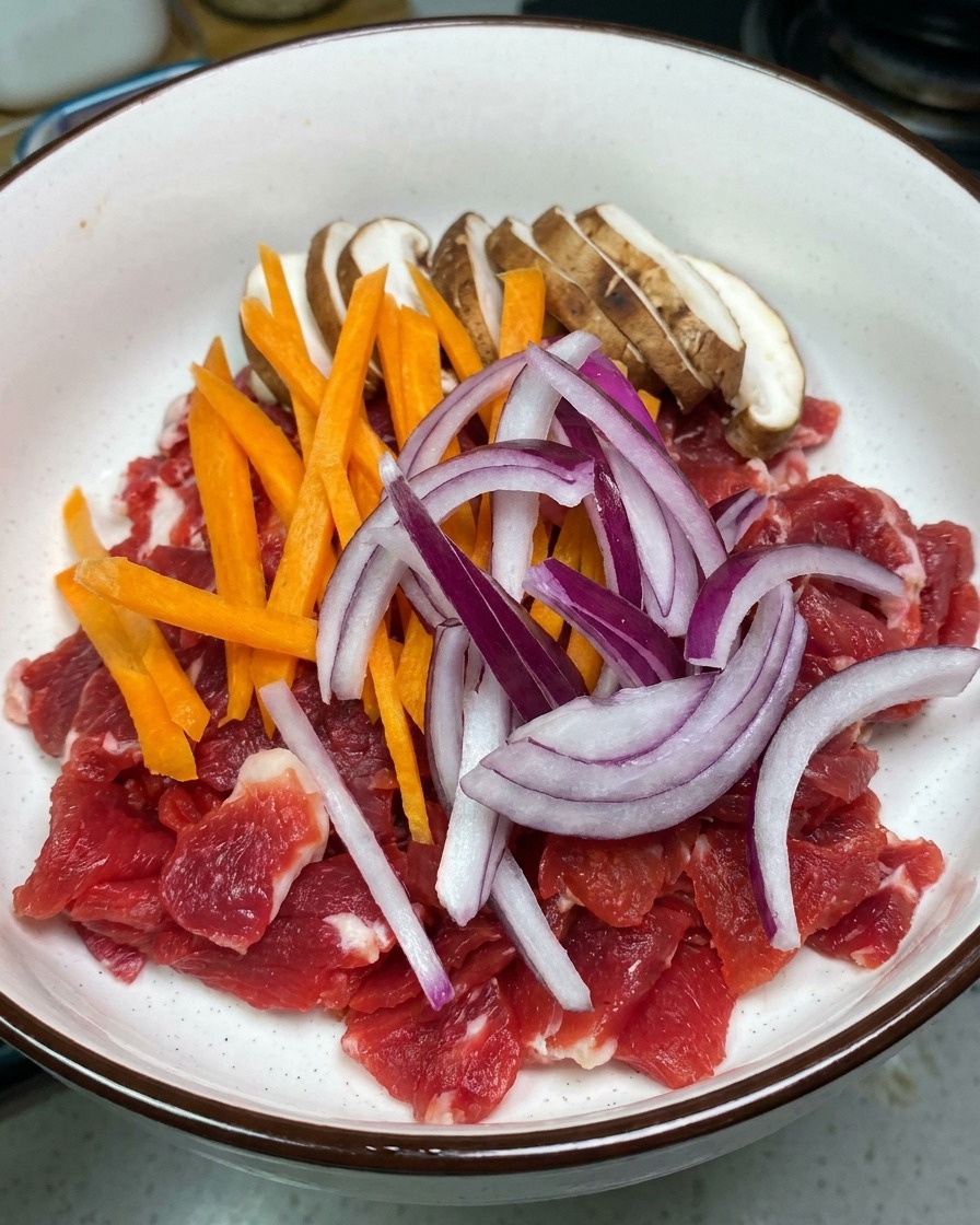 白いボウルに入った薄切りの牛肉の上に、スライスしたしいたけ、細切りの人参、赤玉ねぎがのっている様子。