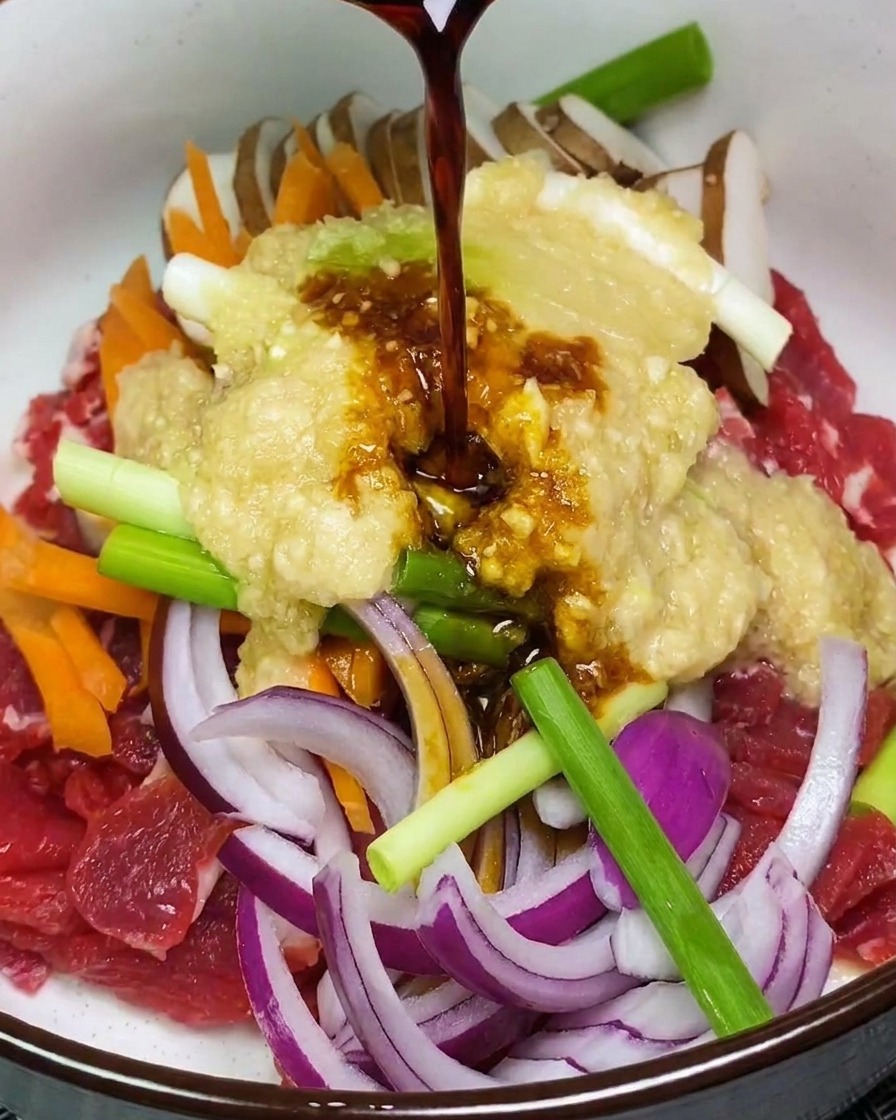 肉と野菜が入ったボウルに、果物ペーストと醤油ベースの濃い色のタレを注いでいる様子。