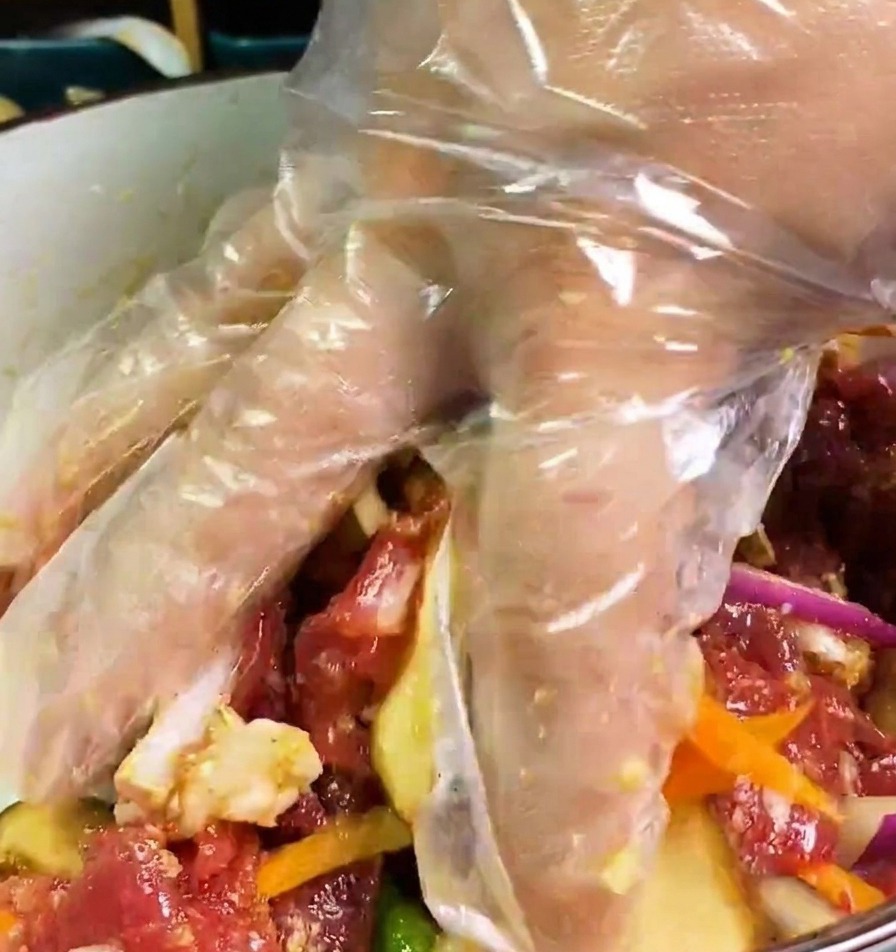ビニール手袋をした手で、ボウルの中の牛肉、玉ねぎ、人参、しいたけをタレと混ぜ合わせている様子。