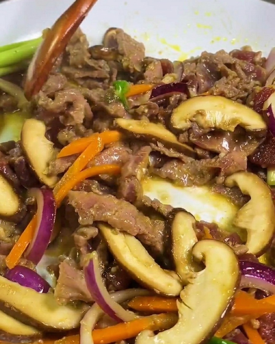 白いフライパンの中で、木べらを使って牛肉、しいたけ、人参を炒めている様子。
