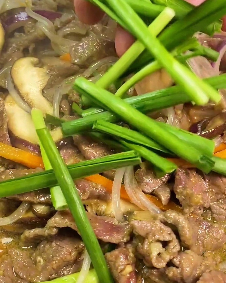 炒めている牛肉と春雨の上に、鮮やかな緑色のカットしたニラを散らしている様子。