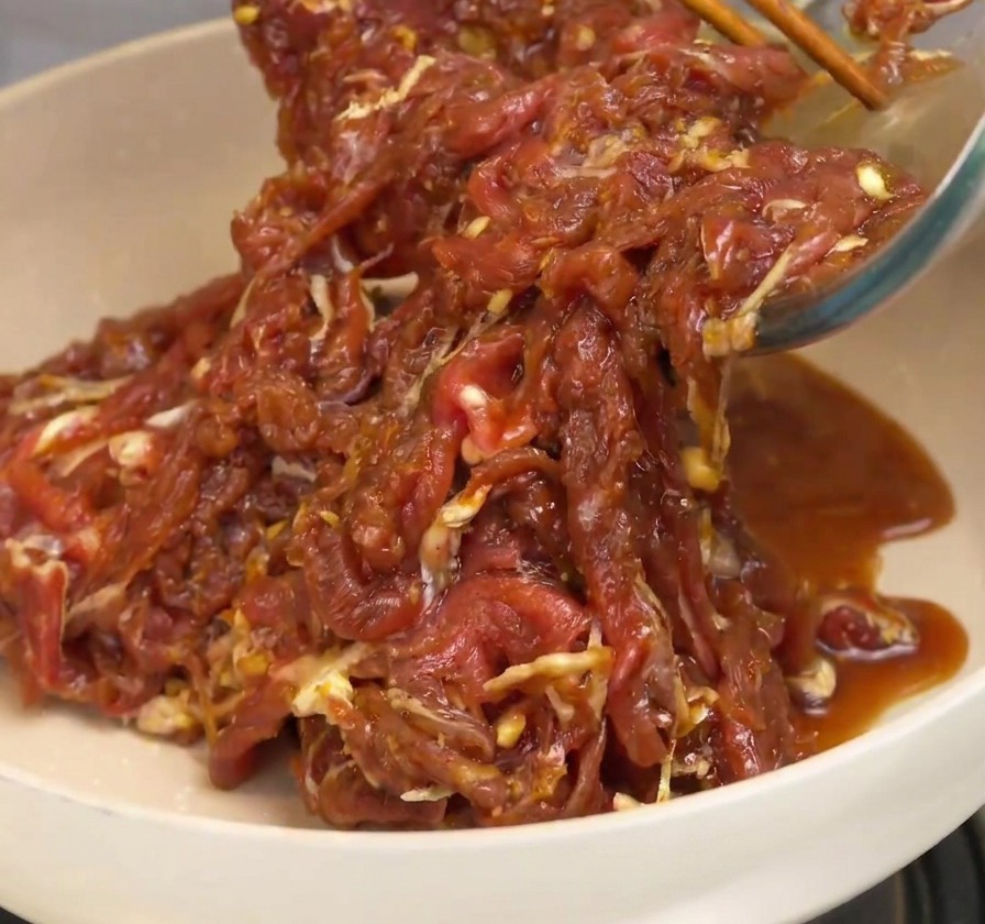 味付けされた牛肉を白いセラミック製のフライパンに入れ、木製のお箸で炒めている様子。