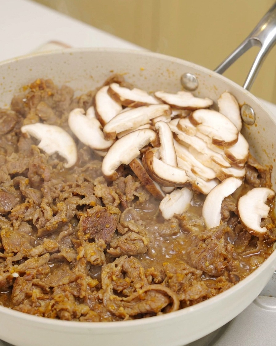 ジュージューと焼ける牛肉の上に、スライスした新鮮な椎茸をのせている様子。